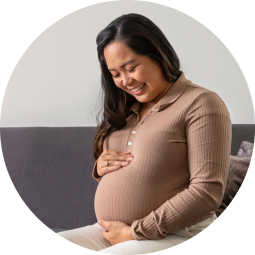 Woman sitting indoors smiling looking at pregnant belly with her hands resting on it